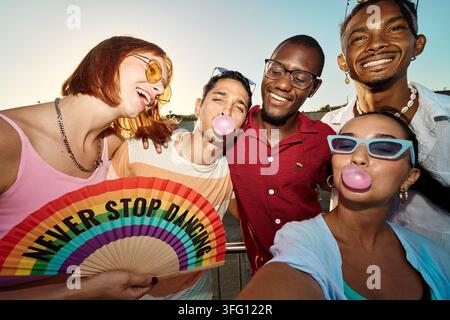 Fröhliche, vielfältige Gruppe von Freunden, die Kaugummi blasen und einen Regenbogenfächer halten, die nie aufhören zu tanzen bei einer Veranstaltung im Freien Stockfoto