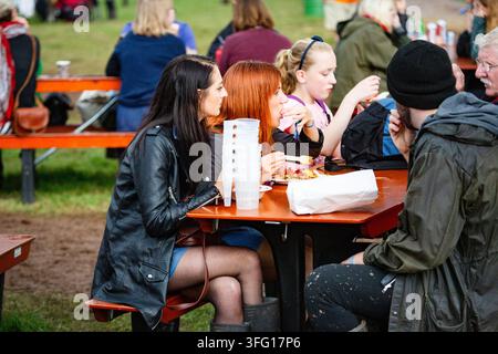 FREUNDE, DIE EINE MAHLZEIT AUF EINEM FESTIVAL TEILEN, REPORTAGE, GREEN MAN FESTIVAL 2015, BRECON, WALES, UK, 2015: Eine Gruppe junger Freunde sitzt zusammen an einem roten Picknicktisch, isst Essen aus Kartons und trinkt aus gestapelten Plastikbechern. Foto: Rob Watkins. INFO: Das Green Man Festival findet jährlich im August in den Brecon Beacons statt. Es feiert alternative Musik, Kunst, Literatur, Wissenschaft und familienfreundliche Kultur in einer wunderschönen Berglandschaft. Das Event 2015 begann sonnig, wurde aber durch nächtlichen Regen an den letzten beiden Tagen schlammig. Stockfoto