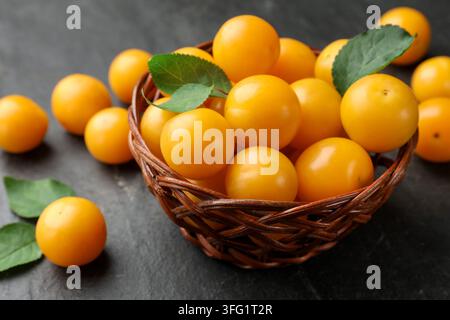 Reife gelbe Kirschpflaumen in einem Weidenkorb auf schwarzem, texturiertem Tisch, Nahaufnahme Stockfoto