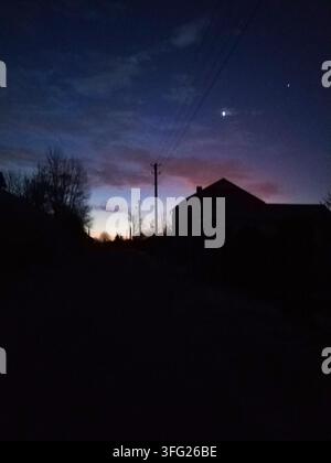 Dunkle Haussilhouetten und Strommasten stehen vor einem tiefblauen Abendhimmel mit hellen Sternen. Die ruhige Dämmerungsstimmung erinnert an friedliche ländliche Einsamkeit Stockfoto