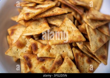 Eine Nahaufnahme, die einen großen Haufen goldbrauner, dreieckiger gebackener Pita-Chips zeigt, die mit Kräutern und Gewürzen gewürzt sind und sich in einer weißen Schüssel befinden. Stockfoto