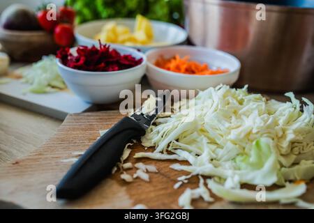 Scheiben Kohl und anderes gehacktes Gemüse sind auf einem Holztisch angeordnet. Schüsseln mit frischen Zutaten stehen in der Nähe, bereit zum Kochen. Stockfoto