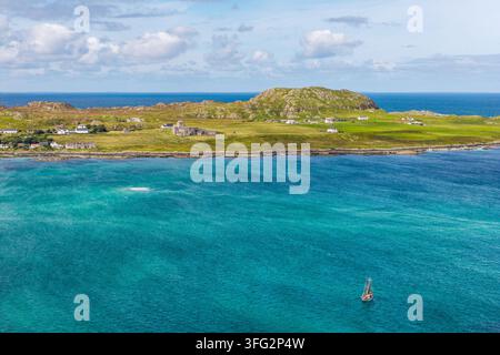 Kleines Holzboot segelt über eine klare blaue Ozeanbucht, abgelegene Küsteninsel-Landschaft mit historischen Steinbauten und felsigen Ufern Stockfoto