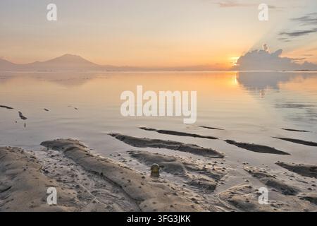 Markante Sandmuster bei Ebbe am Strand von Sanur während eines ruhigen goldenen Sonnenaufgangs, mit der Silhouette des Vulkans Mount Agung und traditionellen Pavillons auf Bali. Stockfoto