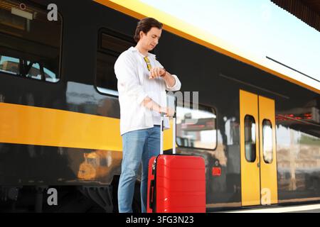 Mann wartet auf einem Bahnhofsgleis, überprüft seine Uhr und hält einen roten Koffer. Reise-, Reise- und Transportkonzepte. Hochwertige p Stockfoto