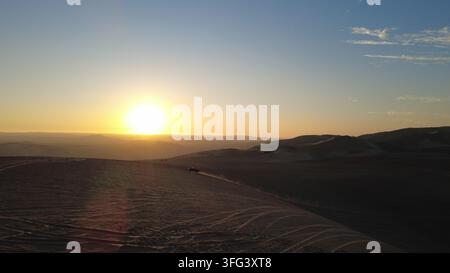 Luftbild-Drohnenaufnahme der Sanddünen von Huacachina bei Sonnenuntergang, Wüste Ica, Peru. Die goldene Sonne geht am Horizont über massiven Dünen-Silhouetten unter. Stockfoto