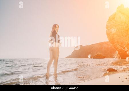 Frauen-Strandurlaub, junge Frau in schwarzem Bodysuit, steht am sandigen Ufer bei Sonnenuntergang mit felsigen Klippen im Hintergrund Stockfoto