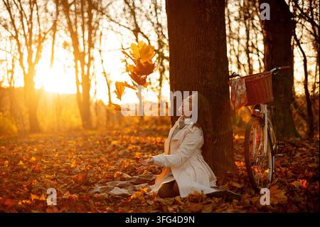 Eine junge Frau lächelt mit geschlossenen Augen und genießt die fallenden Blätter in einem sonnenüberfluteten Herbstwald, während ihr Fahrrad in der Nähe steht Stockfoto