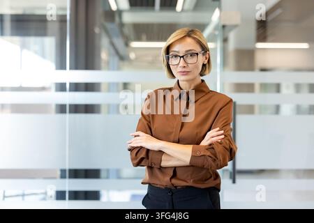 Selbstbewusste Geschäftsfrau posiert mit verschränkten Armen in einer zeitgenössischen Büroumgebung, die Professionalität, Expertise und eine erfolgreiche Karriere in einer Unternehmenskultur repräsentiert Stockfoto