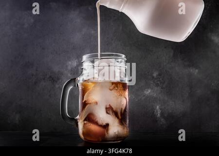 Milch wird aus einem Krug in ein Mason-Glas gegossen, das mit Eiskaffee und Eiswürfeln gefüllt ist, wodurch sich ein Wirbelmuster vor einem dunklen Schieferhintergrund bildet. Stockfoto