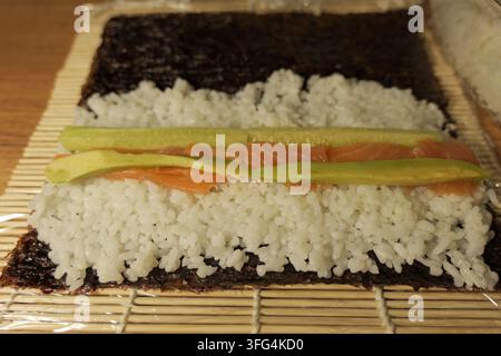 Nori-Algenblatt mit Sushi-Reis, geschnittenem Lachs, Avocado und Gurke auf einer Bambusmatte ausgelegt, bereit zum Rollen von frischem hausgemachtem Maki mit lebendigen Zutaten Stockfoto