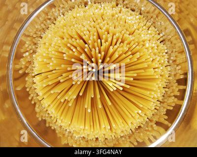 Oberblick-Flachlegung von ungekochter Spaghetti-Pasta, die in einem dichten Sternburst-Kreis in einem glänzenden Metallbehälter arrangiert ist, und zeigt geometrische Essenskunst. Stockfoto
