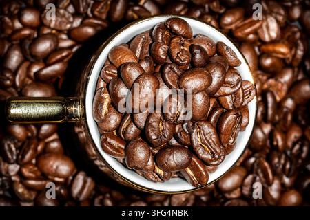 Oberansicht Flat Lay einer weißen Espressotasse mit goldenem Griff, die mit gerösteten Kaffeebohnen überfüllt ist, auf einem Vollbild-Hintergrund aus Kaffeebohnen. Stockfoto