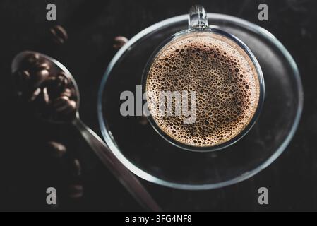 Oberblick-Flachlage eines gläsernen Bechers mit heißem Espresso-Kaffee mit reichen Cremablasen, serviert neben einem Löffel gefüllt mit gerösteten Kaffeebohnen auf einem dunklen Untergrund Stockfoto