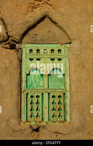Nahaufnahme eines verwitterten grünen Holzfensters mit einem geschnitzten Gitterladen, das in eine rissige Lehmziegelmauer eingelassen ist, Gouvernement Dhofar, Mirbat, Oman Stockfoto