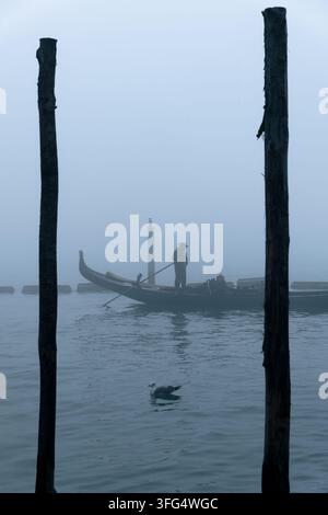 Eine neblige, stimmungsvolle Szene in Venedig mit einer Gondel und einem Gondoliere, eingerahmt von zwei dunklen Holzpfählen (Briccole) im Vordergrund. Stockfoto