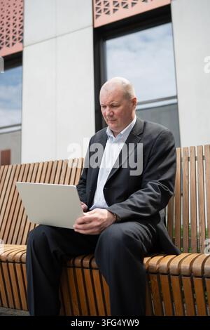 Erfahrener Geschäftsmann arbeitet an einem Laptop, während er auf einer Holzbank vor einem modernen Bürogebäude sitzt, was mobile Arbeit und Geschäftstechnologie repräsentiert. Ver Stockfoto