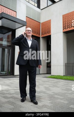 Erfahrener Geschäftsmann in schwarzem Anzug, der draußen vor einem modernen Bürogebäude am Smartphone spricht und Geschäftskommunikation und Unternehmenslebensstil repräsentiert. Ve Stockfoto