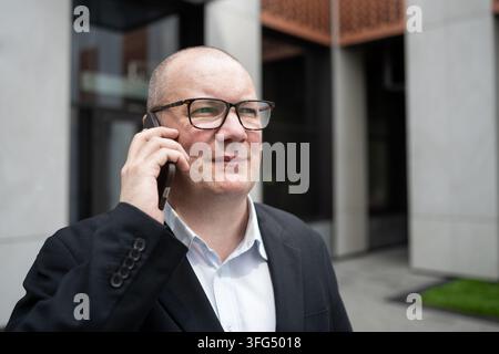 Erfahrener Geschäftsmann mit Brille lächelt, während er draußen vor einem modernen Büro am Smartphone spricht, und repräsentiert positive Geschäftskommunikation und erfolgreichen Ne Stockfoto