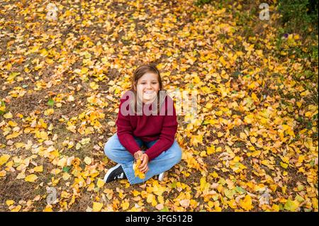 Fröhliches Mädchen, das auf dem mit gelben Blättern bedeckten Boden sitzt Stockfoto