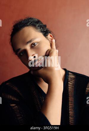 Junger bärtiger Mann mit lockigem Haar, der seine Hand auf die Wange legt, trägt einen schwarzen Strickpullover, Studio-Porträt vor einem warmen Hintergrund Stockfoto
