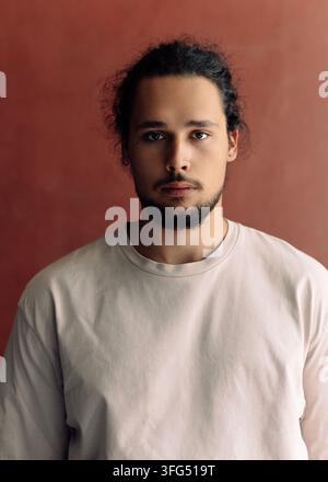 Porträt eines jungen bärtigen Mannes in einem cremefarbenen T-Shirt vor rotem Hintergrund, ruhiger und ernster Ausdruck, Studiofotografie Stockfoto