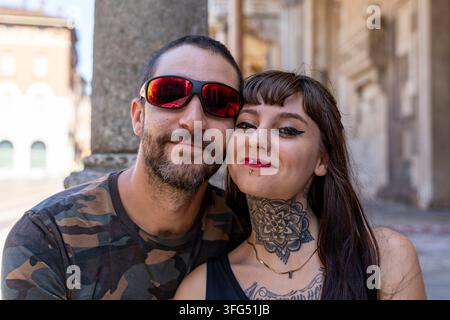 Fröhliches, diverses Paar mit Tattoos und Piercings, das an einem sonnigen Tag im Freien auf einer Stadtstraße zusammen posiert Stockfoto