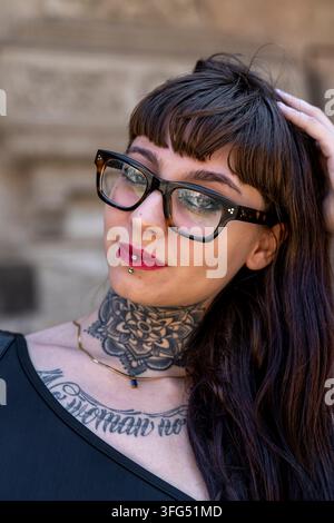 Alternative Frau posiert, zeigt Hals- und Brusttattoos, Lippenpiercing und Brille Stockfoto