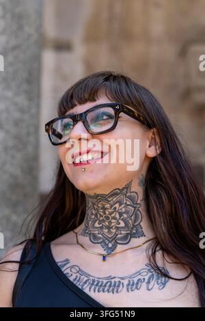 Glückliche junge Frau mit Hals- und Brusttattoos und Piercings lächelt und schaut nach oben draußen Stockfoto
