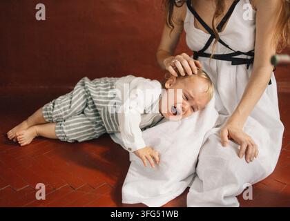 Mutter tröstet ihr weinendes Kleinkind auf dem Boden, sanfter Pflege-Moment, weiches natürliches Licht Stockfoto