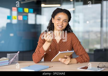 Junge Frau lächelt in die Kamera, hält einen Stift und ein Notizbuch, während sie Geschäftsstrategien plant und Notizen in einer hellen, zeitgemäßen Büroumgebung macht Stockfoto