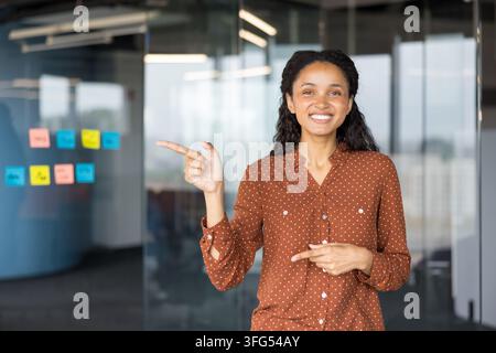Junge Frau steht in einem modernen Büro, lächelt und zeigt mit beiden Händen aktiv auf bunte Merkhinweise an einer Glaswand und zeigt Engagement und Zusammenarbeit in einem professionellen Umfeld Stockfoto