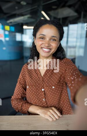 Junge glückliche afroamerikanische Frau, die ihren Arm ausstreckt, um ein Selfie zu machen, selbstbewusst in die Kamera lächelnd, während sie an einem Schreibtisch in einem modernen Büroarbeitsplatz sitzt Stockfoto