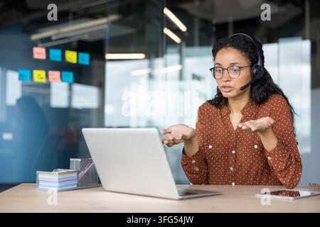 Junge Geschäftsfrau mit Headset, die einen Videoanruf auf einem Laptop führt, arbeitet als Kundensupportmitarbeiterin in einem modernen Büro, bietet Online-Unterstützung und Kommunikation an Stockfoto