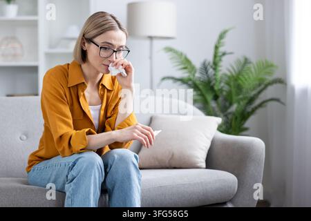 Junge Frau mit Brille sitzt auf einem Sofa zu Hause, wischt sich mit einem Taschentuch die Nase ab und schaut nach unten, einsam und weinend, erlebt Traurigkeit, Trauer und stille emotionale Erschöpfung Stockfoto