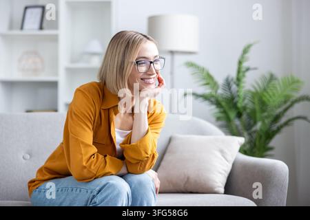 Junge blonde Frau mit Brille, die auf einem bequemen Sofa in einem hellen Wohnzimmer sitzt, lächelt und zur Seite schaut, während sie ihr Kinn auf ihre Hand stützt, und Komfort, Glück und Frieden symbolisiert Stockfoto