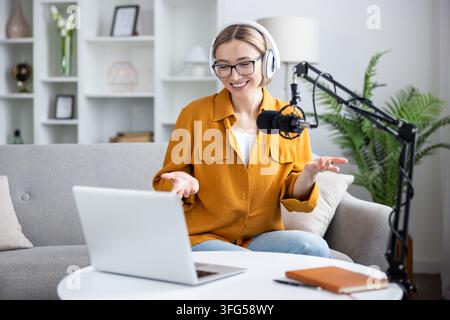 Junge Frau, die einen Podcast erstellt, sendet aus ihrem gemütlichen Heimstudio, trägt Kopfhörer und spricht in ein professionelles Mikrofon mit einem Laptop vor ihr Stockfoto