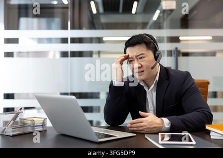Asiatischer Geschäftsmann mit Headset und Anzug, der Kopfschmerzen und Frustration verspürt, während er in einem Büro an einem Laptop arbeitet und während eines Online-Anrufs Herausforderungen und Stress bewältigt Stockfoto