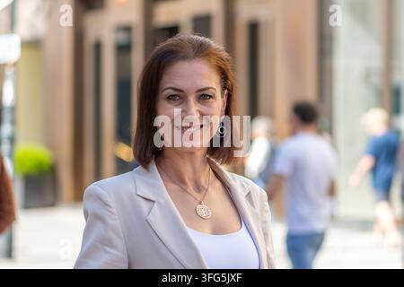 Eine lächelnde Frau mittleren Alters steht im Freien vor einem unscharfen städtischen Hintergrund. Stockfoto
