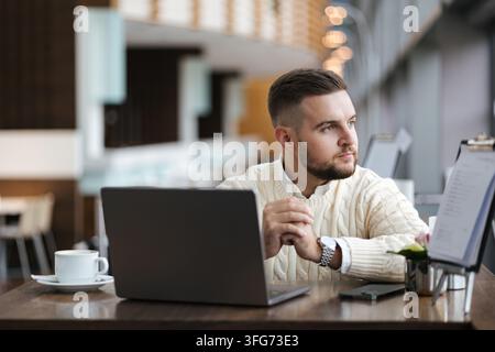 Professionell aussehender junger Mann in einem stylischen Pullover, der am Laptop arbeitet, während er am Telefon sitzt. Moderner Arbeitsbereich mit Tageslicht Stockfoto