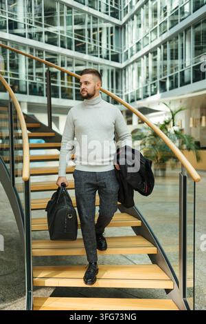 Porträt eines gutaussehenden Mannes im Mantel, der durch das große Büro läuft. Der Kerl hält die Reisetasche. Stilvoller Typ nach dem Arbeitstag Stockfoto