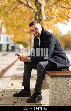 Stylischer Geschäftsmann in einem eleganten Mantel, der im Freien sitzt und an einem Herbsttag telefoniert. Warme Herbstfarben, moderne urbane Umgebung und natürlich Stockfoto