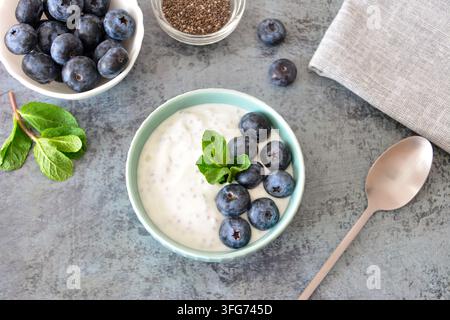 Yoghurt-Schale mit Blaubeeren und grünem Minzblatt auf grauer Oberfläche mit Löffel und Serviette, Flachlayout Stockfoto