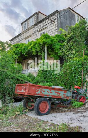 Ein altes Steinhaus in einer mediterranen Umgebung, bedeckt mit üppigen, überwucherten Weinreben, einer verwitterten Veranda und einem rostigen roten Bauernanhänger im Vordergrund Stockfoto