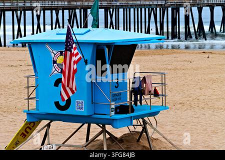 Heller blauer Rettungsturm mit einer amerikanischen Flagge steht auf einem Sandstrand neben Pierpfählen und Ozeanwellen in Pismo Beach, Kalifornien. Stockfoto