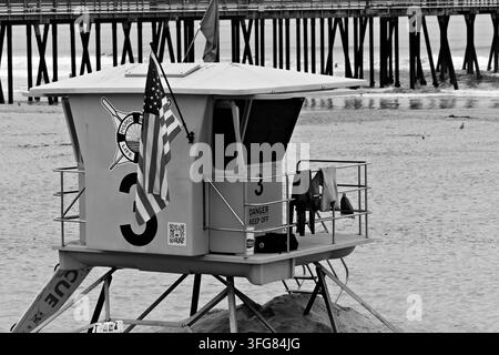 Heller blauer Rettungsturm mit einer amerikanischen Flagge steht auf einem Sandstrand neben Pierpfählen und Ozeanwellen in Pismo Beach, Kalifornien. Stockfoto