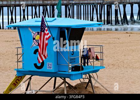 Heller blauer Rettungsturm mit einer amerikanischen Flagge steht auf einem Sandstrand neben Pierpfählen und Ozeanwellen in Pismo Beach, Kalifornien. Stockfoto