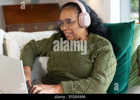 Senior afrikanisch-amerikanische Frau lächelt, während sie auf der Couch zu Hause mit Laptop und rosa Kopfhörern tippt. Wohnzimmer, Homeoffice, Tageslicht, Interieur, gemütlich, Stockfoto