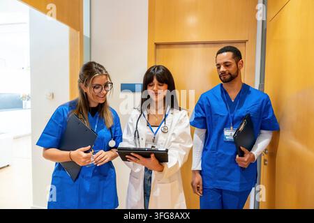 Verschiedene medizinische Fachkräfte in OP-Kleidung und einem Laborkittel arbeiten zusammen und besprechen Patientendaten auf einem digitalen Tablet in einer modernen Klinik oder Ho Stockfoto
