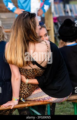 ZWEI FREUNDE UMGARMEN SICH AUF EINEM FESTIVAL, LEIBE KUNST UND TANZ, GREEN MAN FESTIVAL, BRECON, WALES, UK: Zwei junge Frauen teilen eine liebevolle Umarmung, während sie zusammen auf einer Bank bei einem Open-Air-Festival sitzen, von hinten gesehen. Eine große Gruppe von Familien nimmt an einem riesigen Spiel von Musical Chairs teil, gemischt mit einem Tanzwettbewerb. Foto: Rob Watkins. INFO: Das Green Man Festival findet jährlich im Brecon Beacons im August statt. Es feiert Musik, Kunst, Literatur, Wissenschaft und familienfreundliche Kultur in einer wunderschönen Berglandschaft. Umherziehende Künstler tragen zur Atmosphäre auf den Feldern zwischen den Bühnen bei Stockfoto